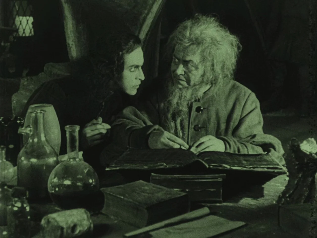 The Golem (1920) movie still: medium — A dimly lit room filled with books and various glass vials on a table. A younger man watches intently as an…