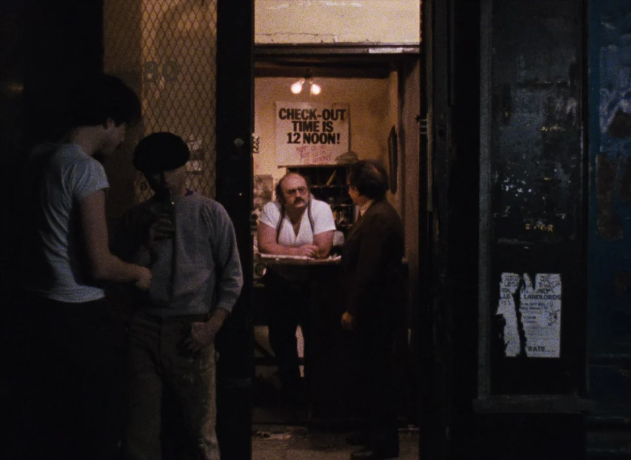 Basket Case (1982) movie still: medium — A dimly lit street entrance reveals a small room with a check-out sign. Three men stand near the doorway…
