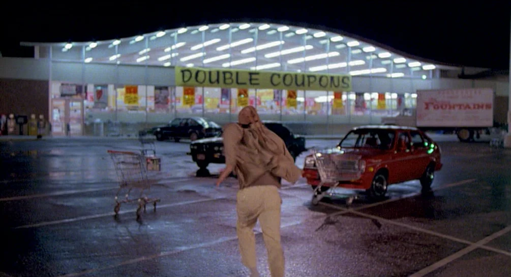 Raising Arizona (1987) movie still: fullbody — A nighttime grocery store parking lot features a large arched facade and neon signage. A man in light…