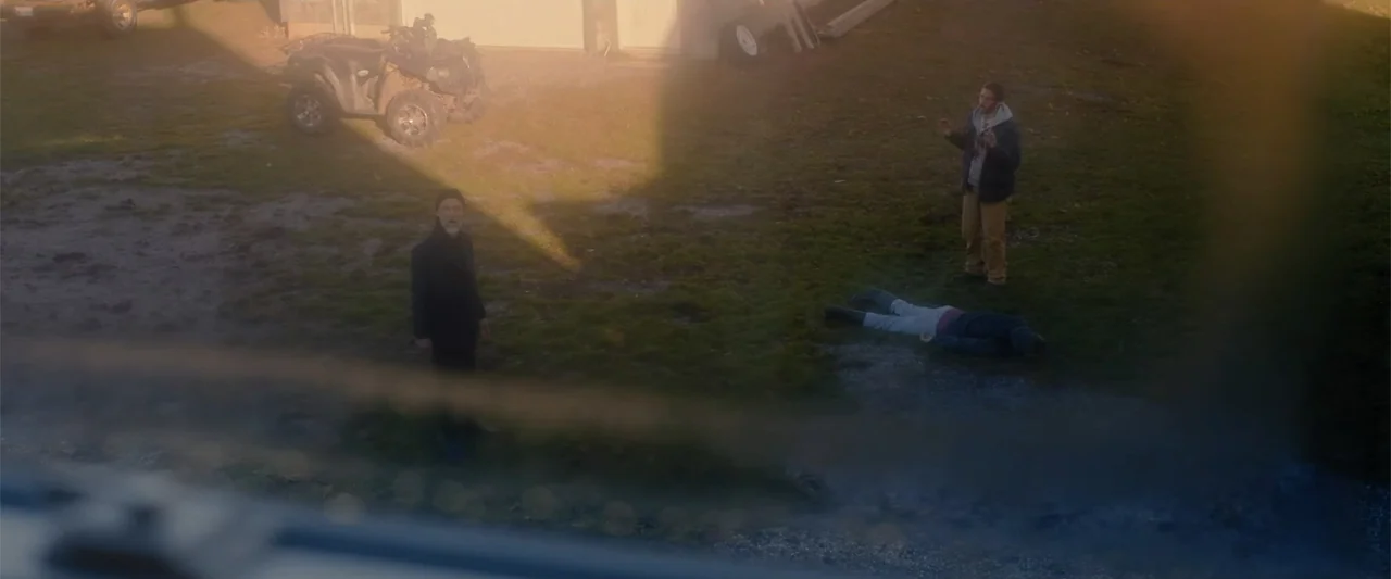A Small Fortune (2021) movie still: wide — A grassy yard features an all-terrain vehicle and a figure prone on the ground. Two men remain standing…