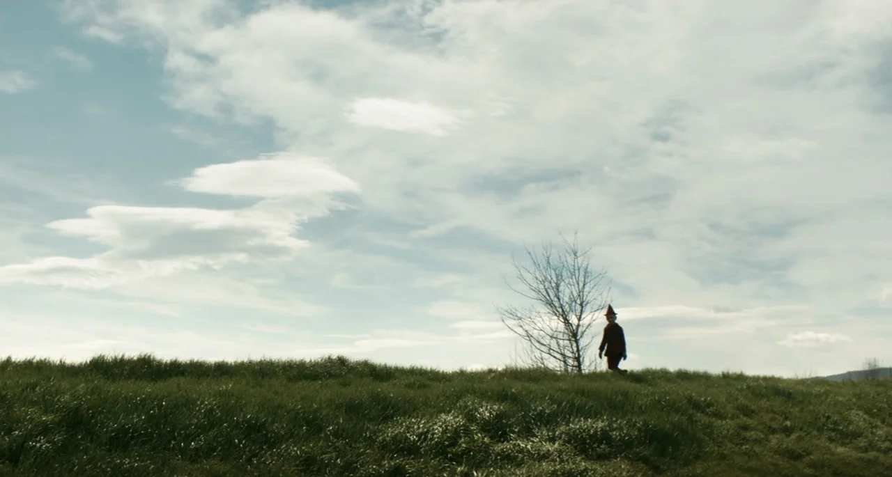 Pinocchio (2022) movie still: wide — An open field of green grass slopes upward toward a bright cloudy sky. A small figure in a dark costume walks…