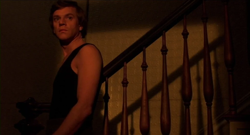 Cat People (1982) movie still: medium — An interior staircase with wooden spindles and textured wallpaper is lit by dim warm light. A young man in a…