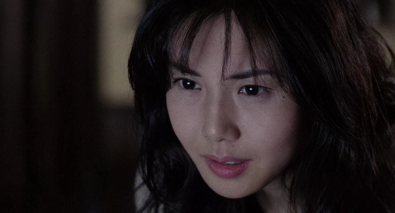 Ringu (1998) movie still: closeup — A dimly lit interior space with an indistinct background in deep shadow. A young woman with dark hair looks…