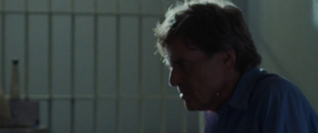 The Old Man & The Gun (2018) movie still: medium — A dimly lit room features a set of vertical metal bars in the background. An older man sits in…