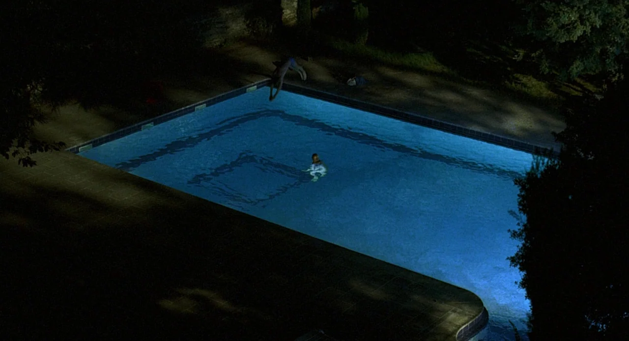 Swimming Pool (2003) movie still: wide — A rectangular pool is centered in a dark residential backyard at night. One person leaps into the water…