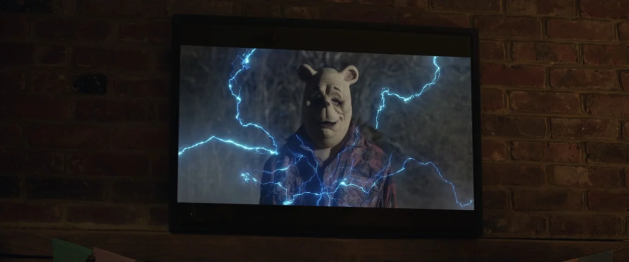 Winnie-the-Pooh: Blood and Honey 2 (2024) movie still: medium — A television screen displays a character wearing a bear mask amidst animated blue…