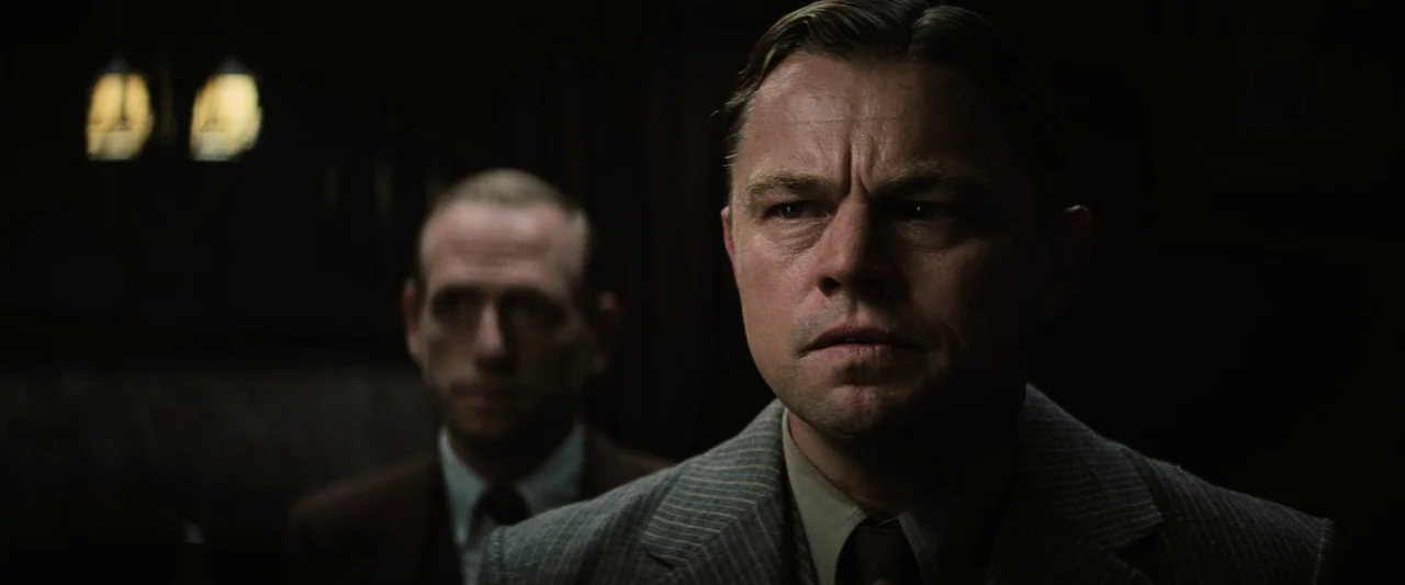 Killers of the Flower Moon (2023) movie still: medium — A dark, dimly lit room with wood paneling and indistinct architectural details. One man…