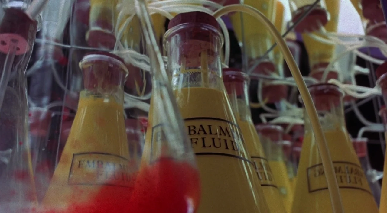 Dr. Phibes Rises Again (1972) movie still: closeup — A collection of glass flasks filled with opaque yellow liquid sits on a rack. Clear plastic…