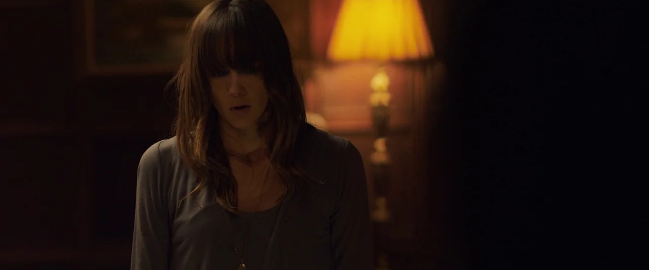 You’re Next (2013) movie still: medium — A dimly lit room with dark wooden walls and a soft glowing table lamp. A young woman with bangs stares down…