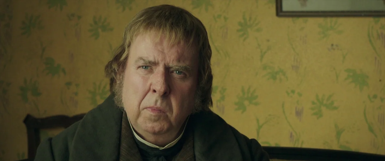Mr. Turner (2014) movie still: closeup — A man stands in front of yellow wallpaper with a repeating green floral pattern. The man with sideburns…