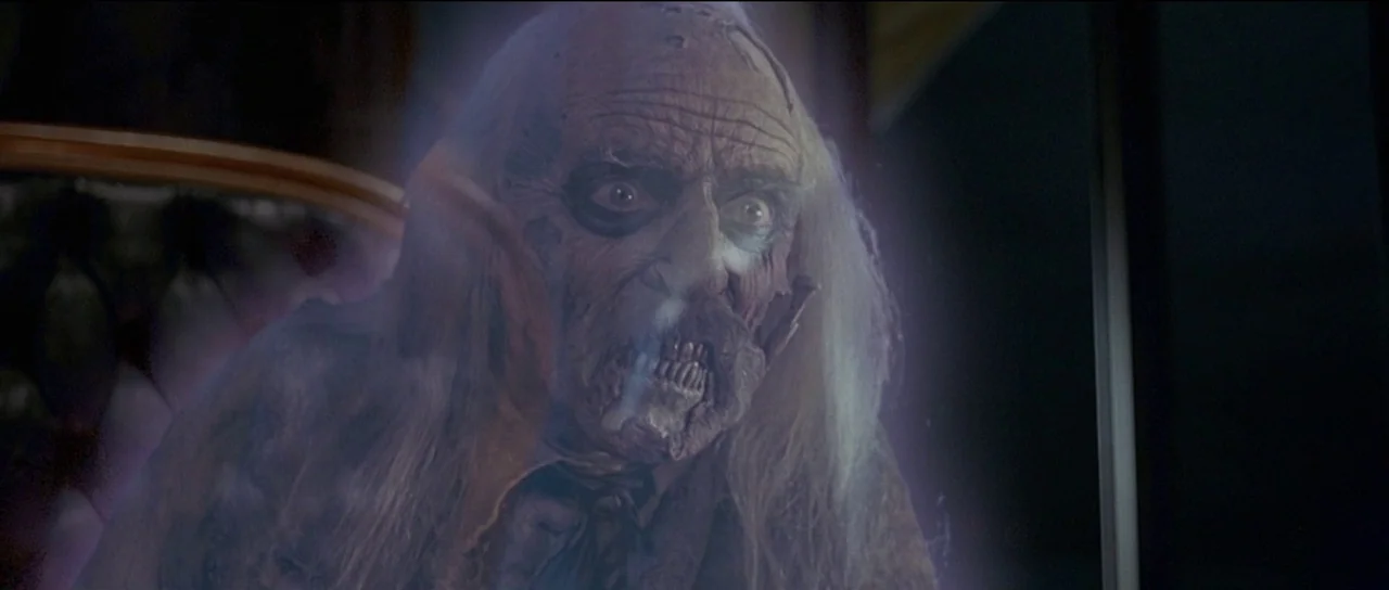 The Frighteners (1996) movie still: closeup — An aged, skeletal figure with pale, weathered skin is centered within a dark space. The subject stares…