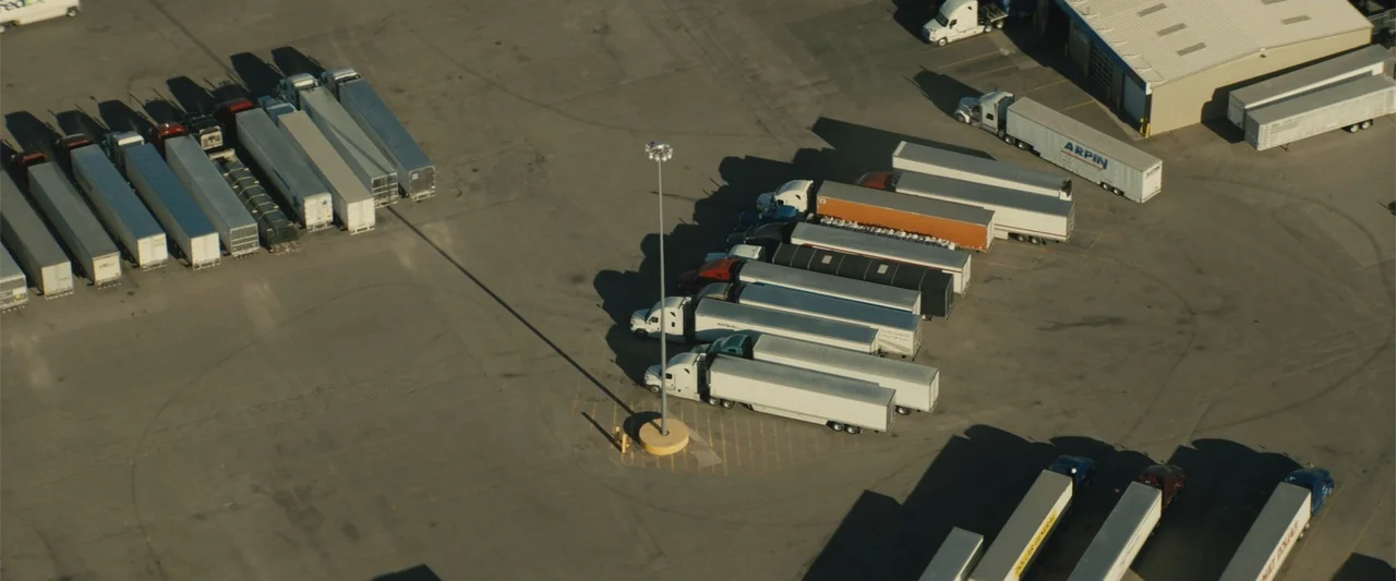 Those Who Wish Me Dead (2021) movie still: establishing — A vast paved trucking terminal contains multiple rows of parked semi-trailer trucks. Long…