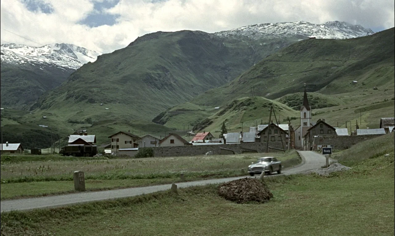 Goldfinger (1964) movie still: wide — A rural Alpine village is nestled in a valley beneath snow-capped mountain peaks. A silver car approaches the…