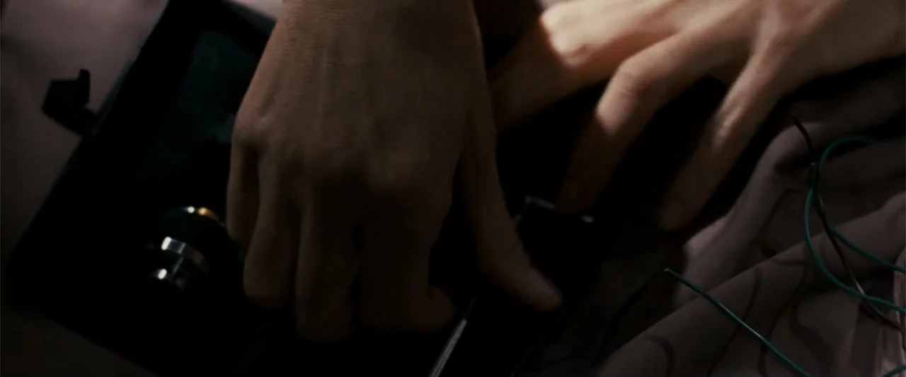 Angels & Demons (2009) movie still: closeup — A close view shows two hands manipulating a black electronic component on a flat surface. Several thin…