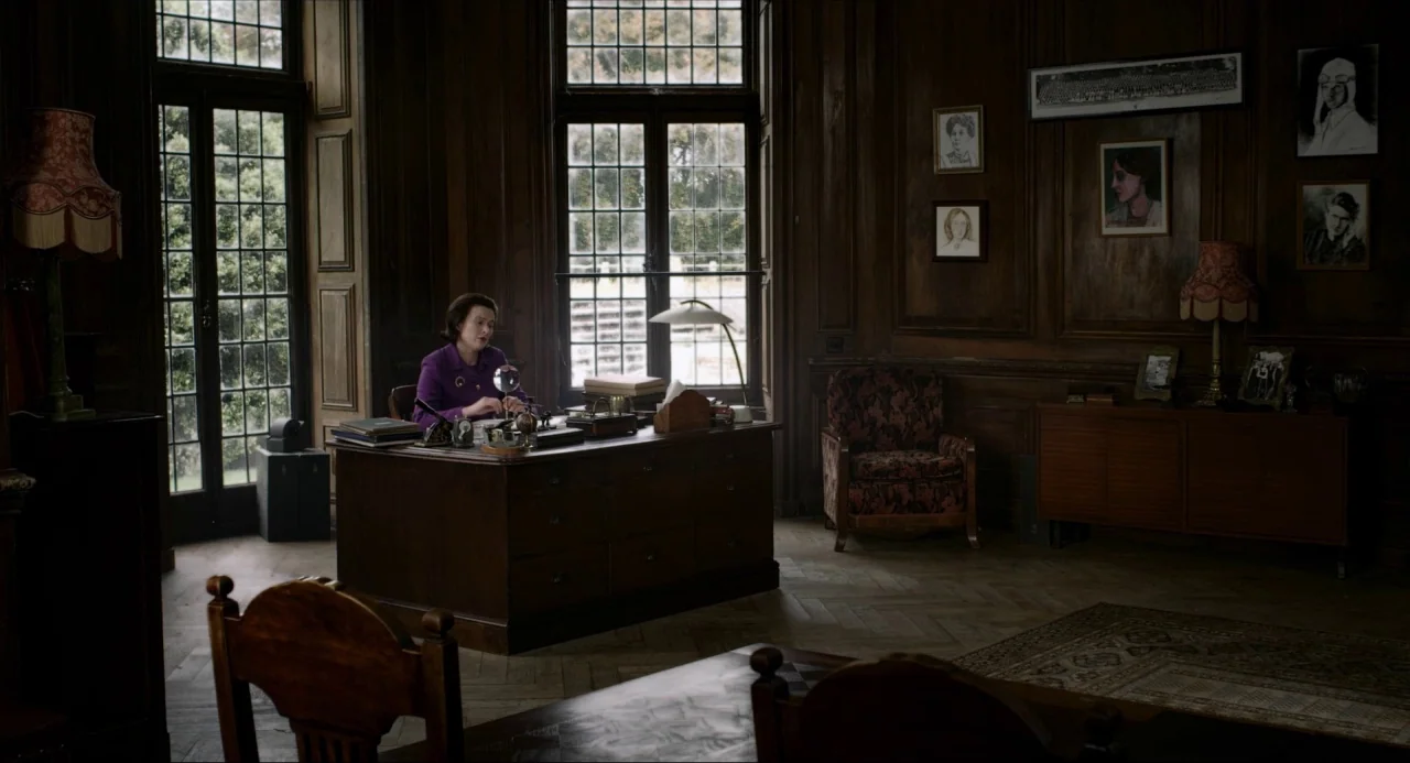 The Falling movie still: wide — A wood-paneled room features high windows, antique furniture, and portraits hung on the walls. A lone woman works at…