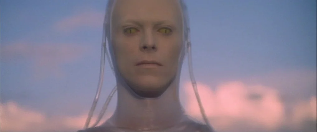 The Man Who Fell To Earth (1976) movie still: closeup — A pale, hairless humanoid figure stands outdoors against a twilight sky. The character…