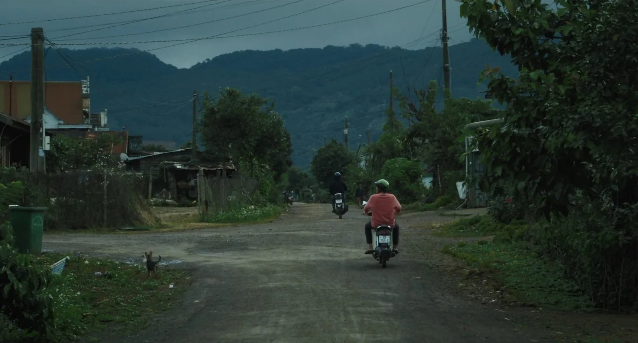 Inside The Yellow Cocoon Shell (2023) movie still: wide — A rural dirt road stretches toward distant mountains under a heavy overcast sky. Two…
