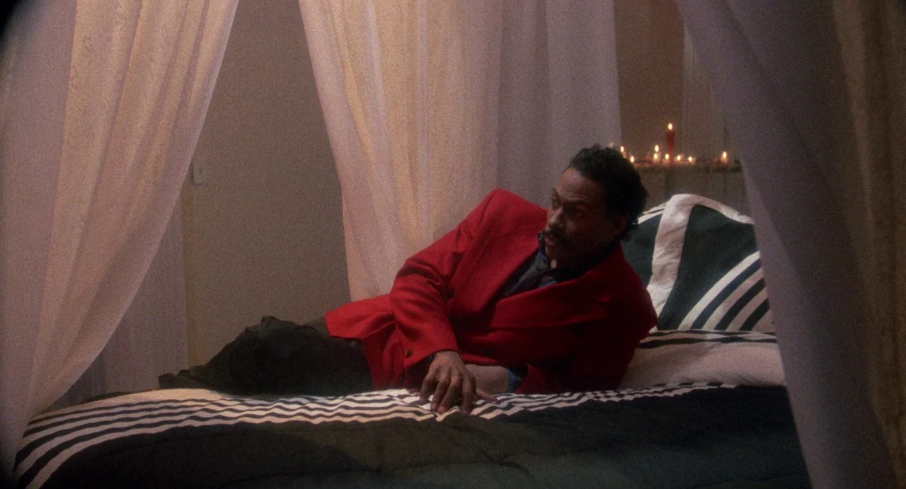 Def By Temptation (1990) movie still: medium — A man lies on a bed with white fabric curtains draping around the frame. He wears a red blazer and…