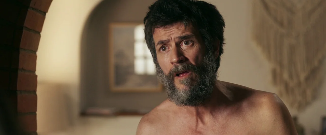 When Evil Lurks (2023) movie still: closeup — A man stands in a room with white walls and a brick archway. He faces forward while shirtless with his…