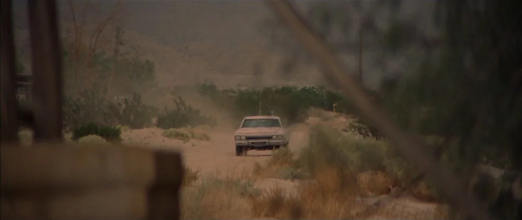 3 Women (1977) movie still: wide — A dusty desert road stretches towards distant hills covered in low-lying arid vegetation. A light-colored sedan…
