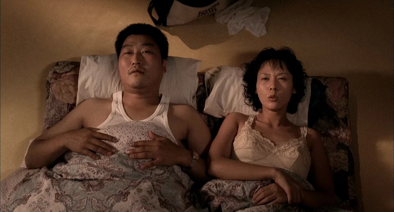 Memories of Murder (2003) movie still: medium — A man and a woman lie side-by-side in bed under a patterned floral quilt. They both stare upward…