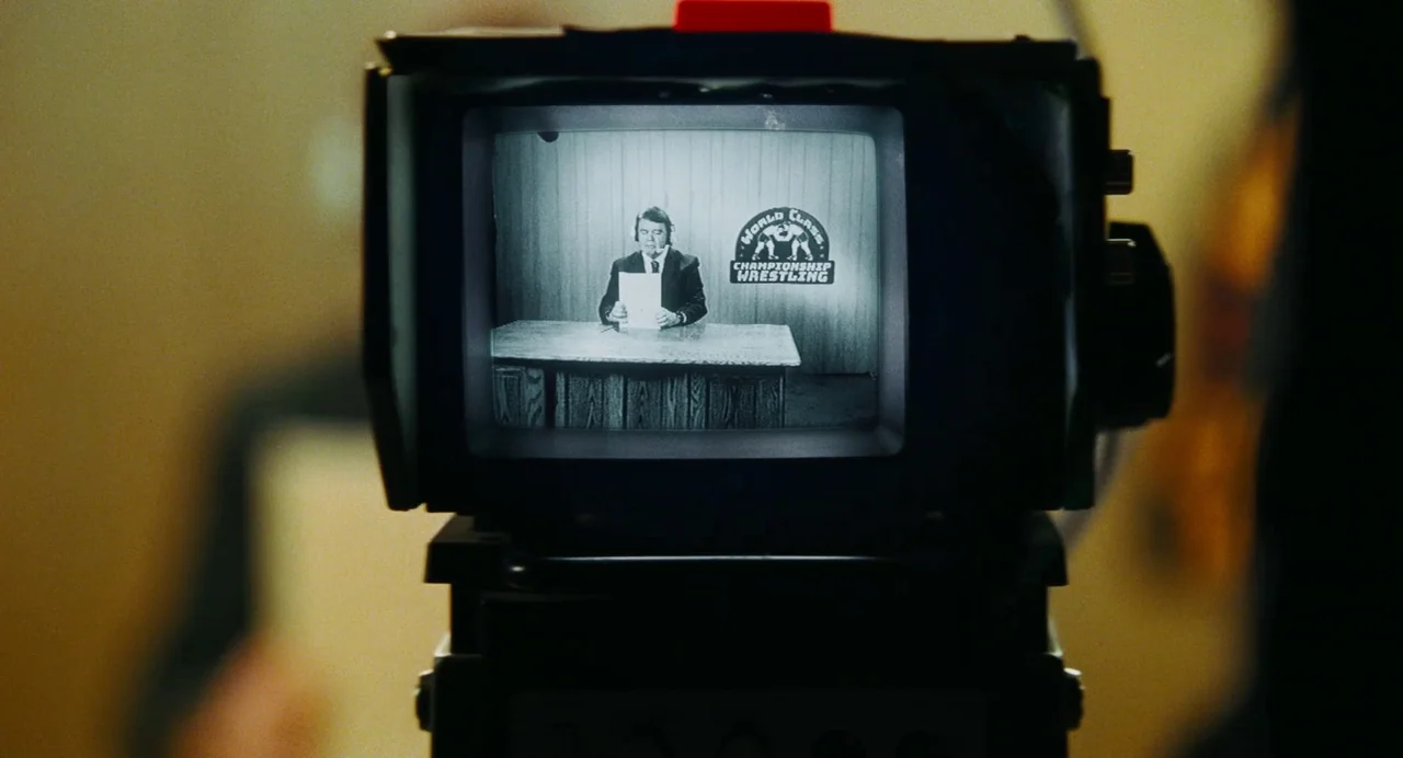 The Iron Claw (2023) movie still: medium — A man in a suit sits behind a desk inside a television news set. He holds a document in his hands while a…