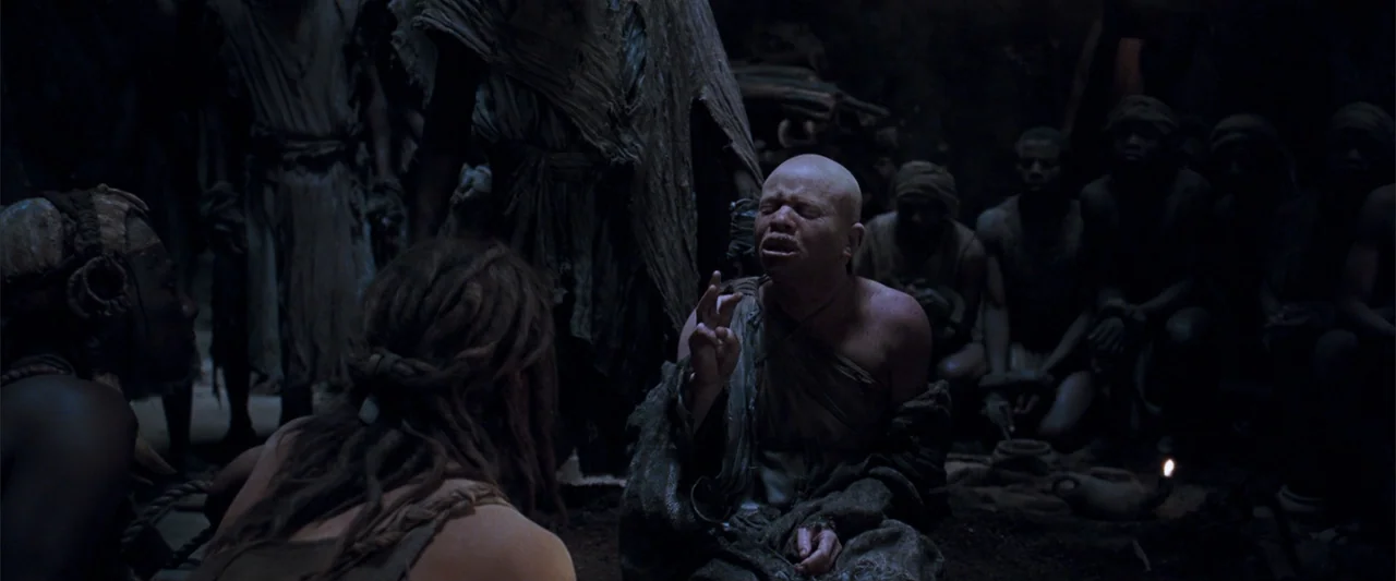 10,000 BC (2008) movie still: medium — A dark, earth-toned interior space with rough stone walls and dim firelight illumination. A group of people…