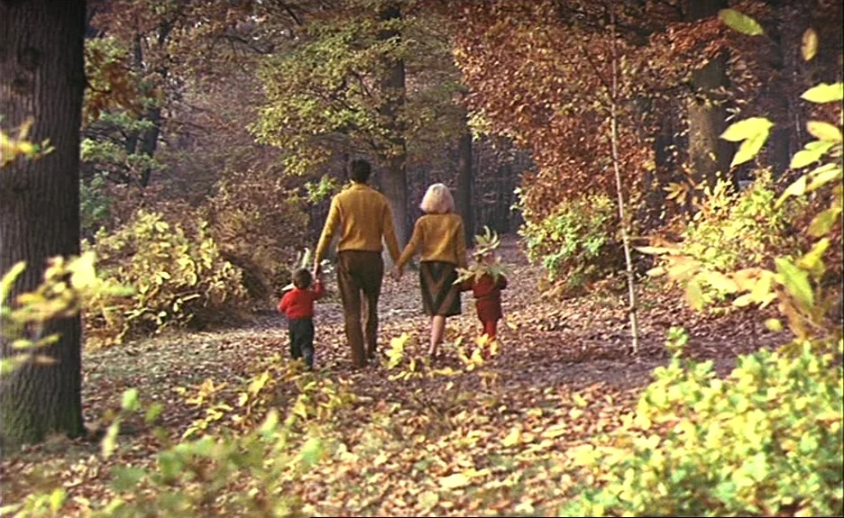 Le Bonheur (Happiness) (1965) movie still: fullbody — A dense autumn woodland with a dirt path covered in fallen brown leaves. A family of four…