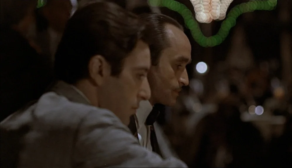 The Godfather Part II (1974) movie still: medium — A dimly lit indoor space with blurred green and white festive lights in background. Two men in…