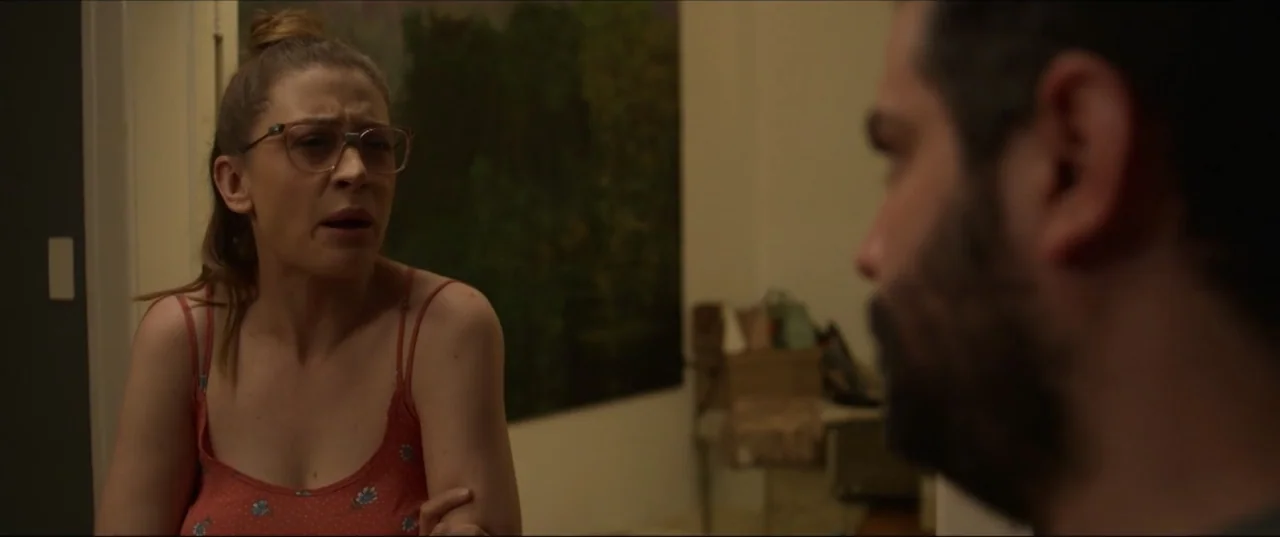 Can't Quit Mom (2022) movie still: medium — A dimly lit room features a woman speaking to a man in the foreground. The woman faces the man while…