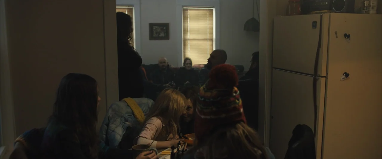 Skin (2009) movie still: medium — A dimly lit apartment room contains a group of people in the background. A child sits at a table in the foreground…