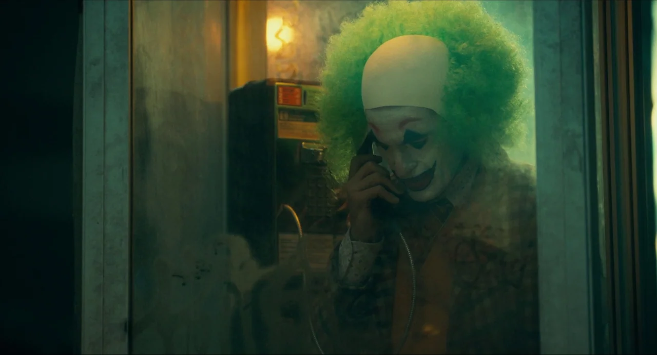 Joker (2019) movie still: medium — A man in clown makeup stands inside a dimly lit outdoor glass phone booth. The green-lit booth is at night and…
