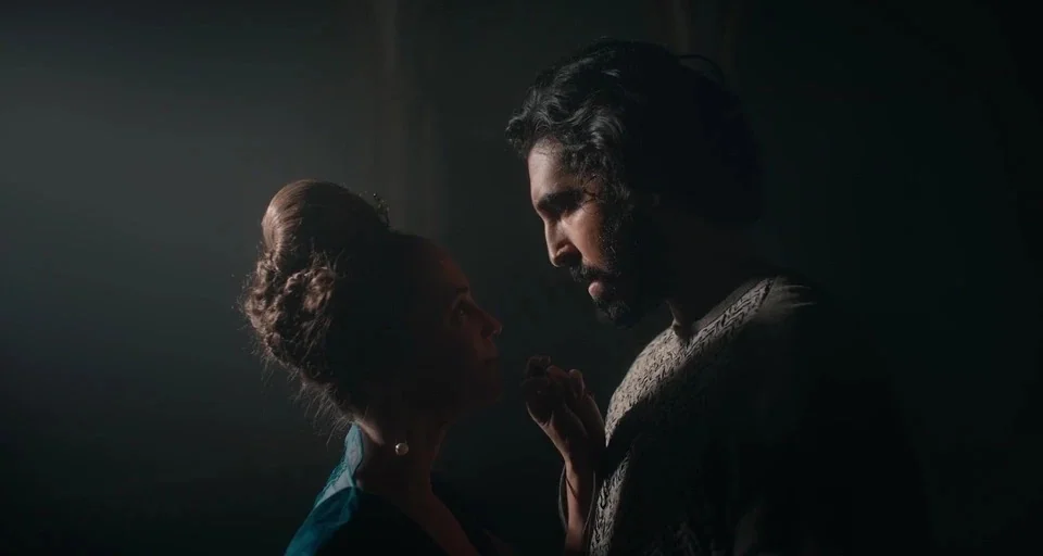The Green Knight (2021) movie still: medium — A dark, dimly lit chamber with stone architecture in a fantasy period setting. A man and a woman stand…