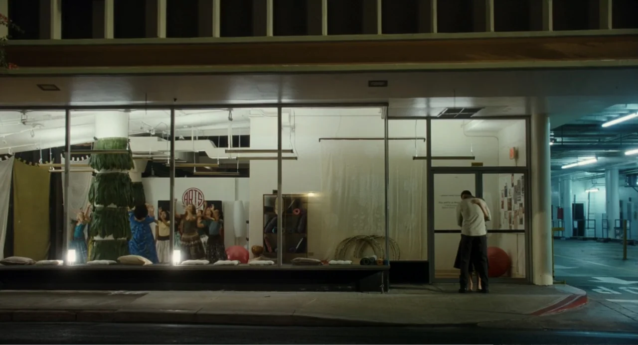 Aloha (2015) movie still: wide — A glass-fronted dance studio at night sits adjacent to a dimly lit concrete parking structure. A man and a woman…