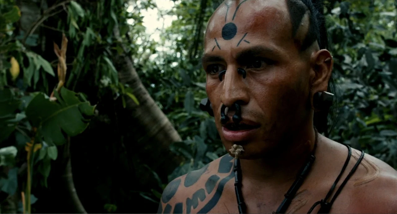 Apocalypto (2006) movie still: closeup — A dense green tropical jungle surrounds a man with facial markings and ornaments. The man stands with a…