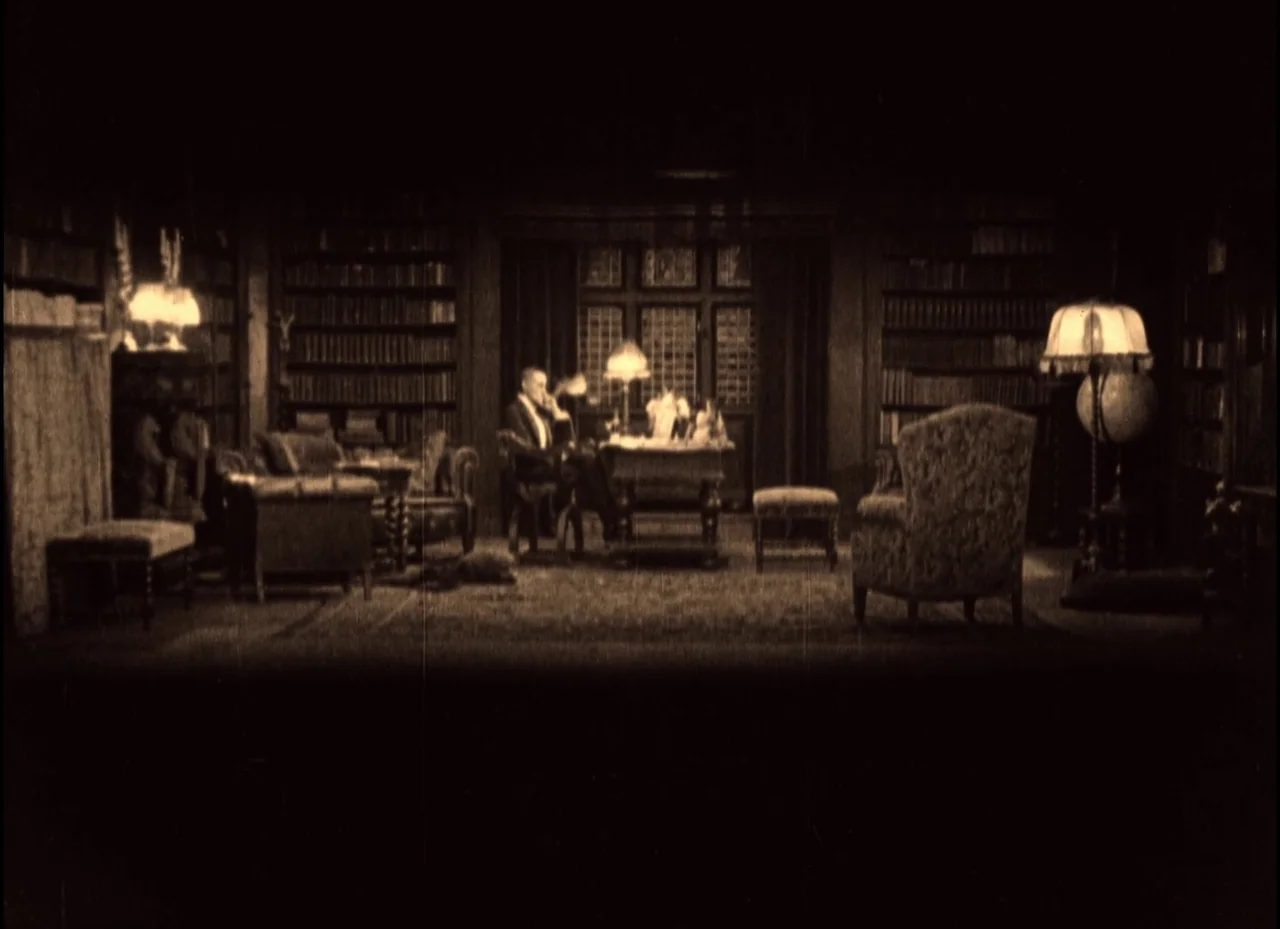 The Phantom Carriage (1921) movie still: wide — A spacious library with tall dark wood bookshelves spanning every wall. A lone man sits at a central…