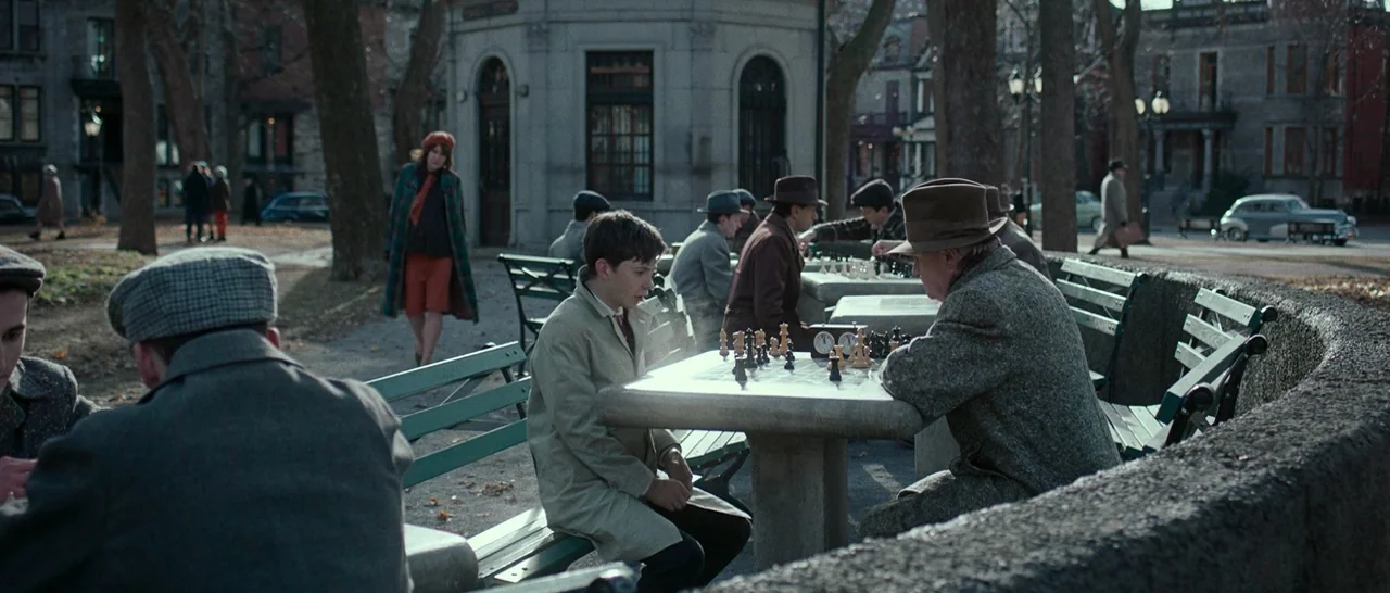 Pawn Sacrifice (2014) movie still: medium — A public park features stone chess tables surrounded by trees and vintage buildings. A teenage boy faces…