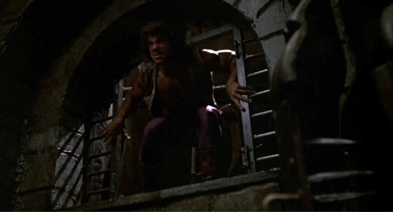 Hercules (1983) movie still: medium — A stone chamber features a heavy metal-barred window opening to darkness outside. A man crouching in the…