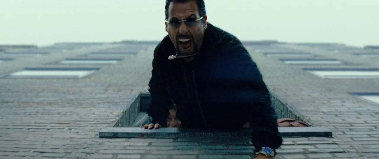 Uncut Gems (2019) movie still: medium — A man leans forward from an open window set into a grey brick building wall. He is shouting with his mouth…