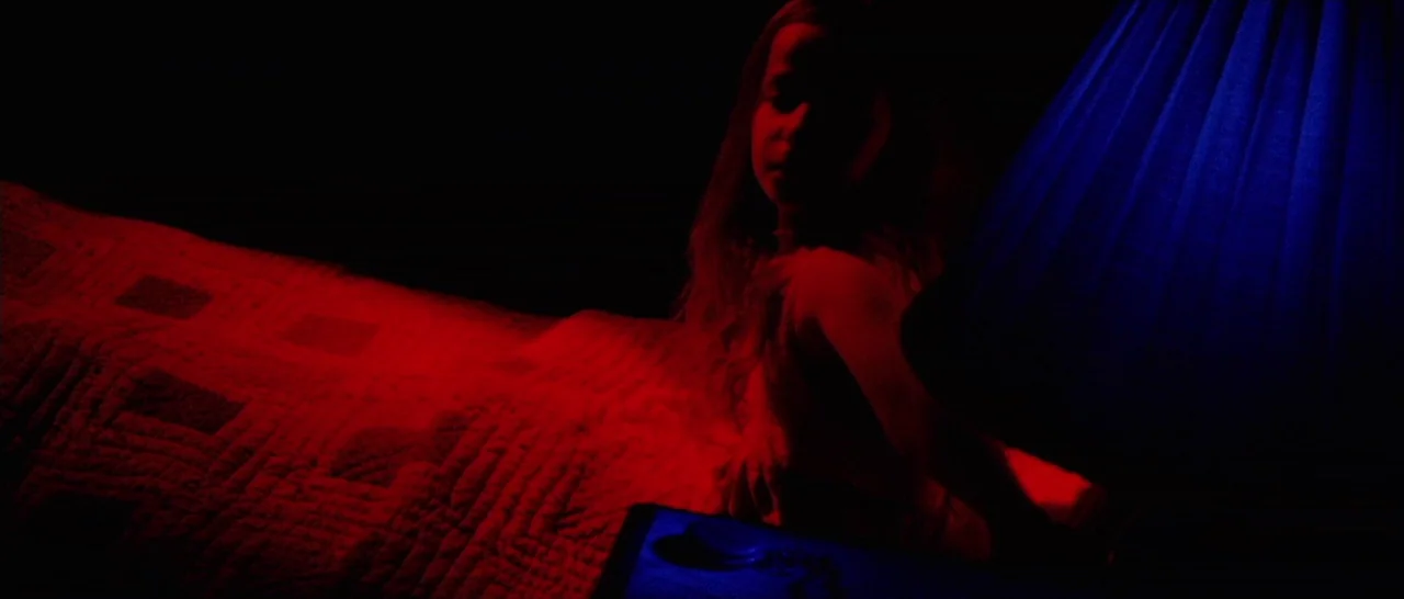 Amer (2009) movie still: medium — A dark bedroom features a bed with a textured quilt illuminated in red. A young girl sits on the edge of the bed…