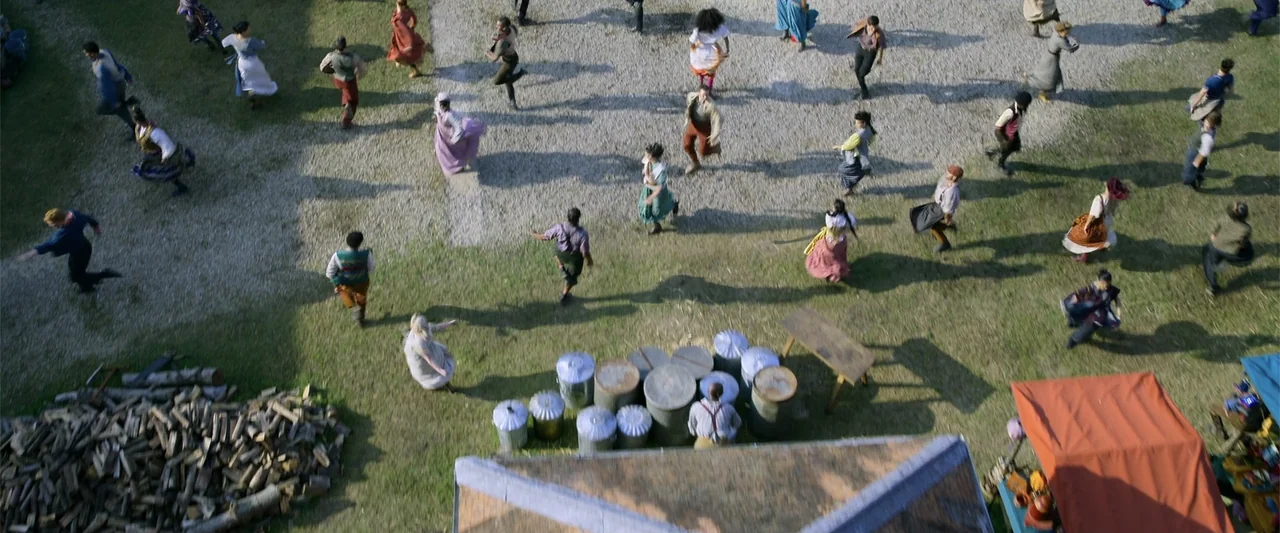 Cinderella (2015) movie still: wide — A crowd of people runs across a grassy field dotted with gravel and metal barrels. The scene is viewed from an…