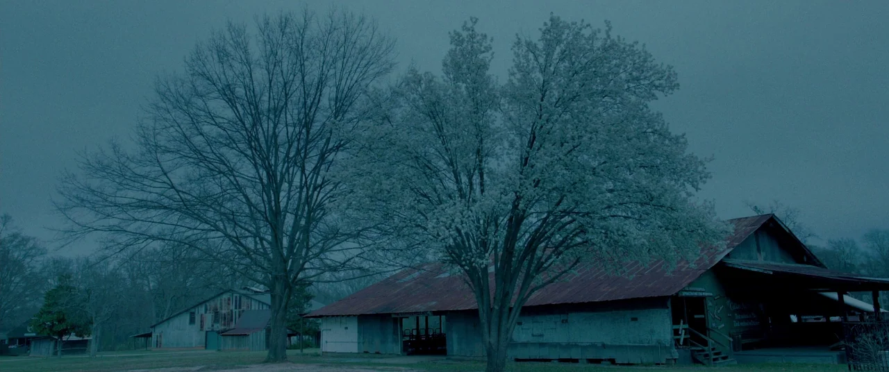 Lawless (2012) movie still: establishing — A rural farm landscape features old wooden buildings with rusted metal roofs under overcast skies. Two…