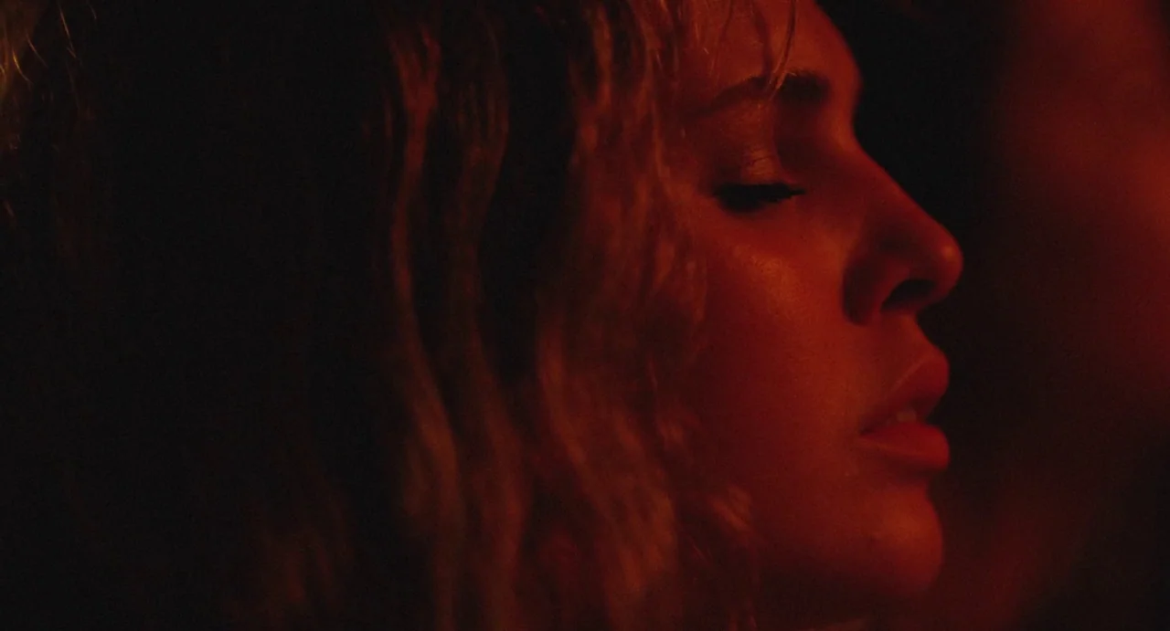 The Giant (2019) movie still: closeup — A woman's face is illuminated by a deep red light in a dark space. She holds her eyes closed with her head…