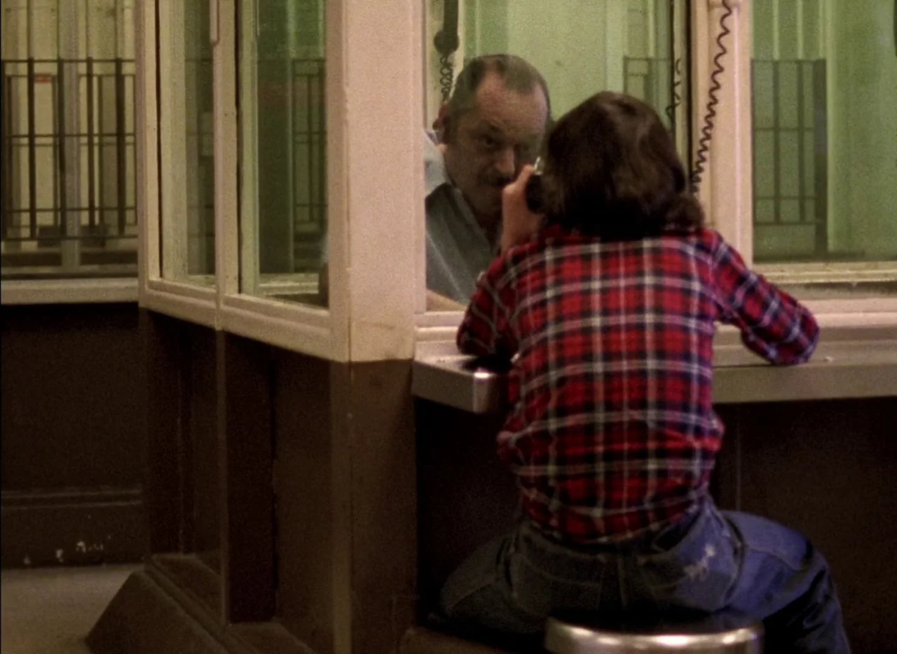 Streetwise (1984) movie still: medium — A utilitarian visitation booth features thick glass windows and a metal counter surface. A man in a red…