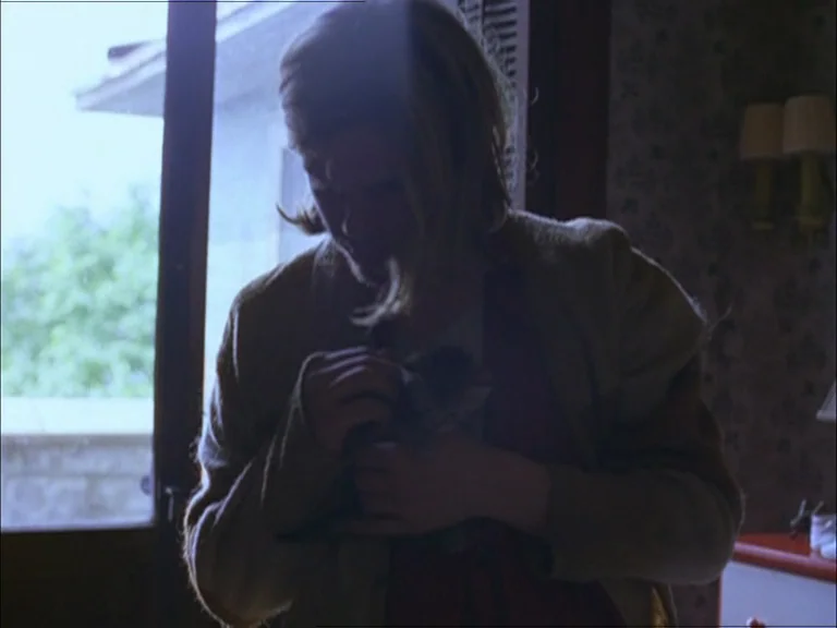 Last Days (2005) movie still: medium — A dimly lit room features a window with an view of an outdoor ledge. A person with blonde hair stands holding…