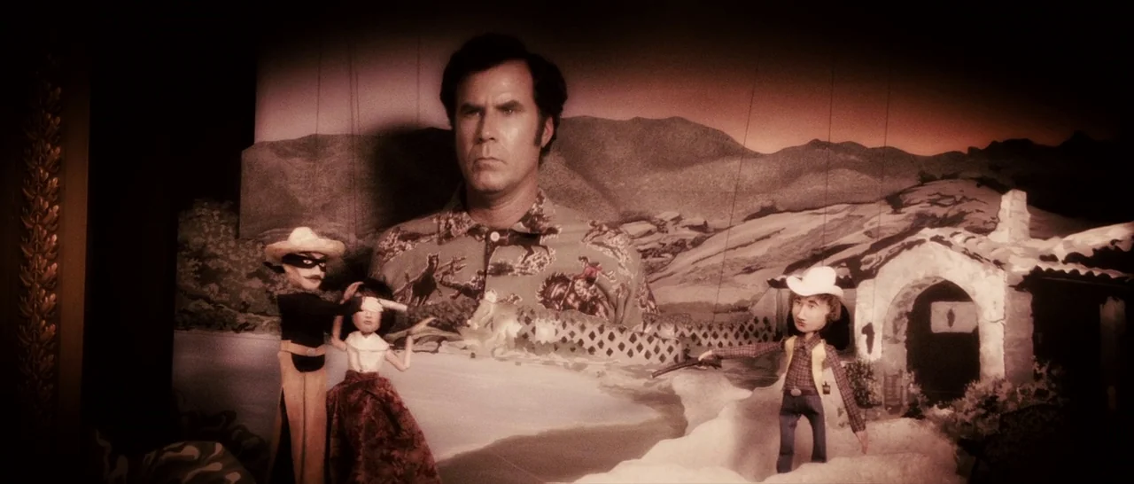 Casa De Mi Padre (2012) movie still: medium — A man stands behind a miniature desert puppet stage. The puppets are arranged in front of his torso.