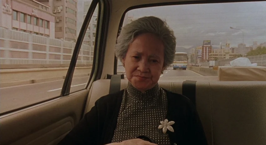 Yi-Yi (A One and A Two) (2000) movie still: medium — An elderly woman sits in the back of a moving car during the day. Urban buildings and a highway…