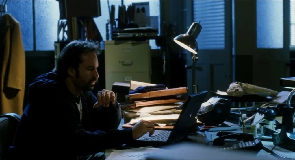 Narc (2002) movie still: medium — A dimly lit workspace is filled with stacks of paperwork and filing cabinets. A man works at a laptop while blue…