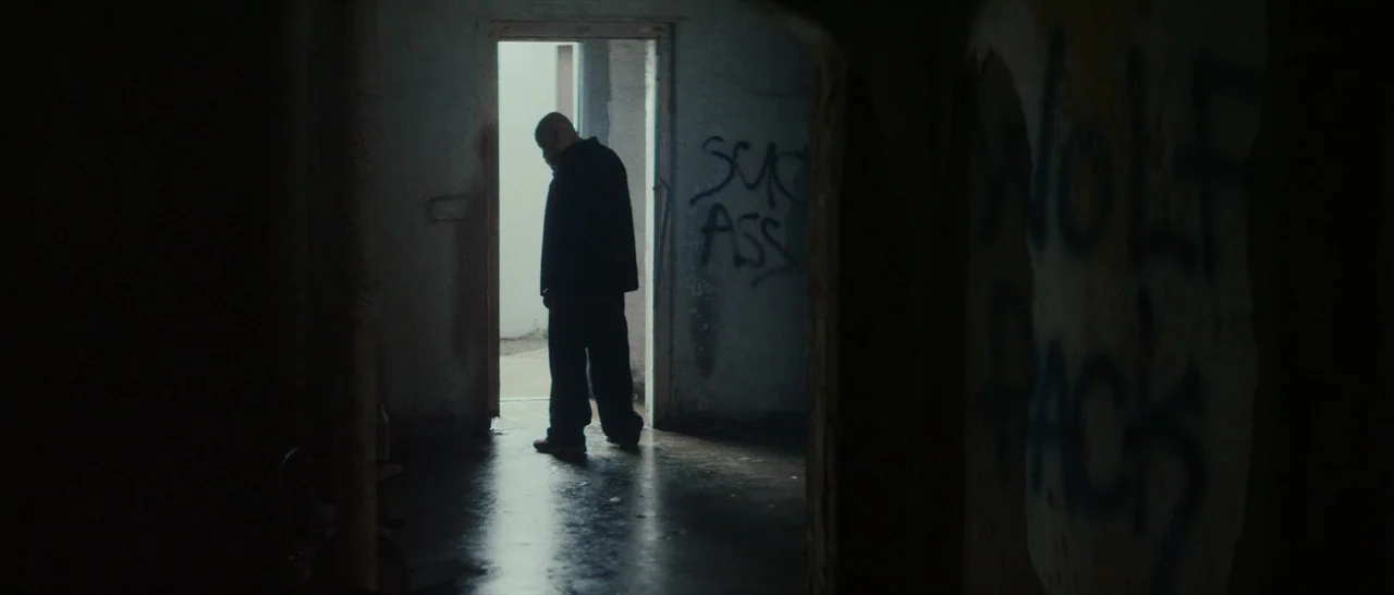 Dog Eat Dog (2016) movie still: medium — An abandoned interior room with graffiti-covered walls and a concrete floor. A solitary man stands in the…