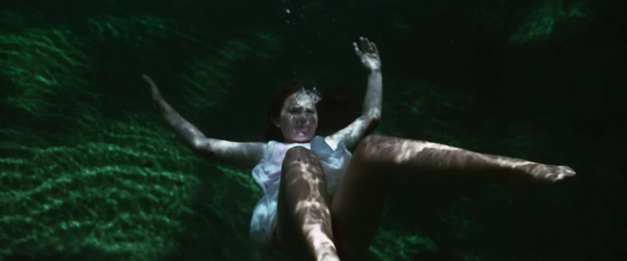 The Mad Hatter (2021) movie still: medium — A person floats underwater in a dark, murky, natural body of water. The subject hangs suspended in the…